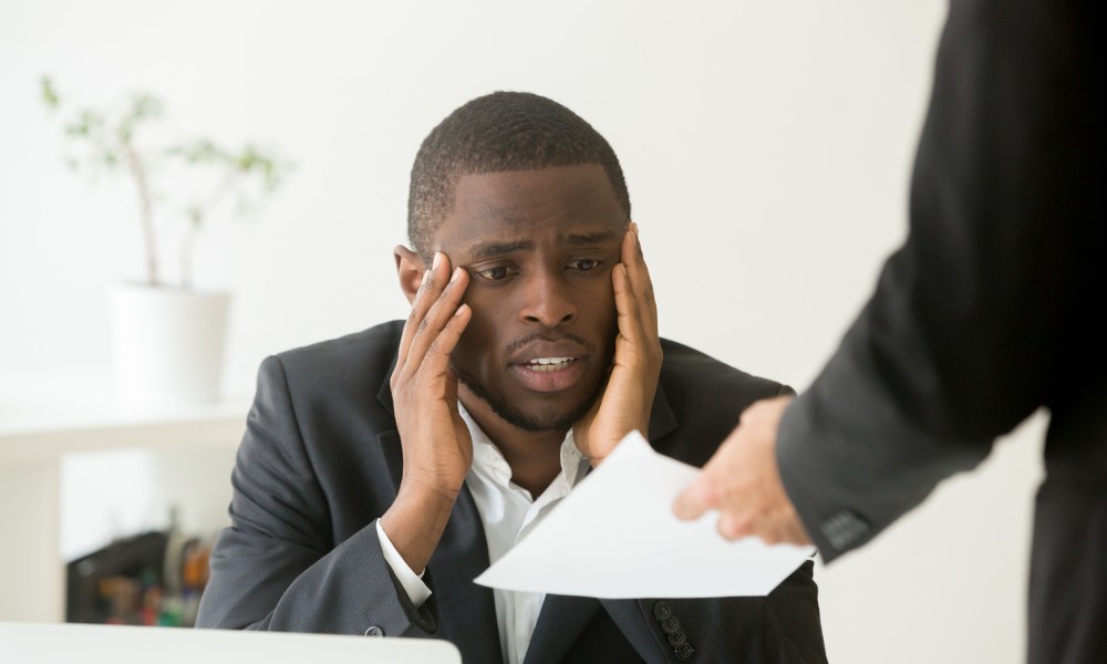 A man at work with his hands on his face looking upset while his boss hands him a sheet of paper.