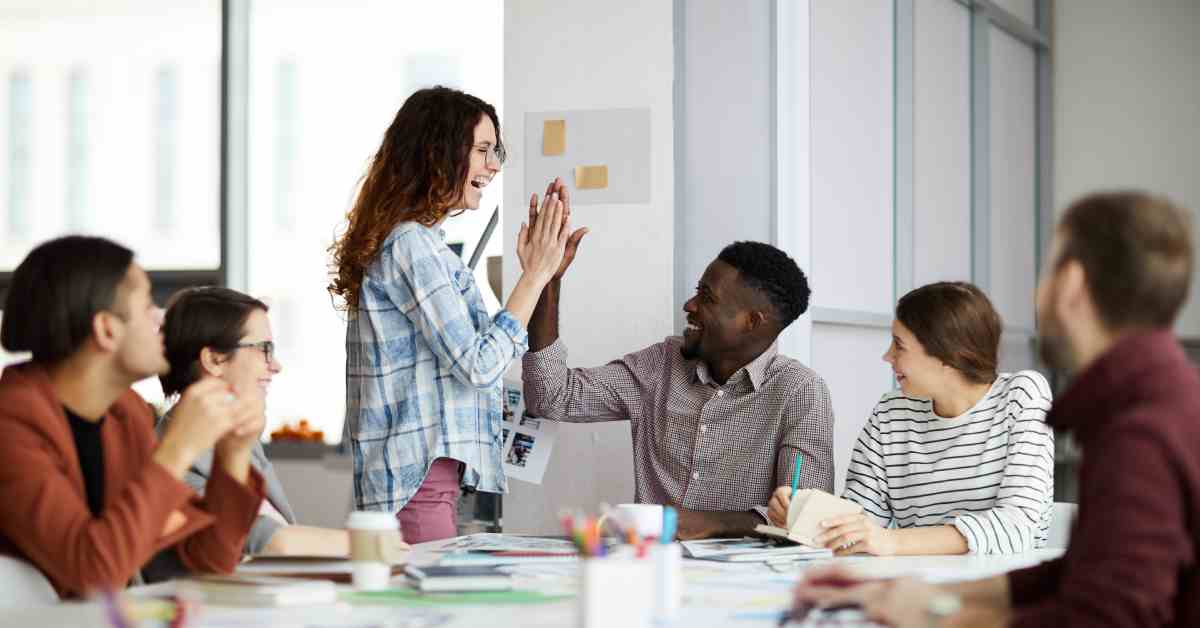 A team of semi-casually dressed employees having a meeting around a table with one of the employees happily high-fiving the other.