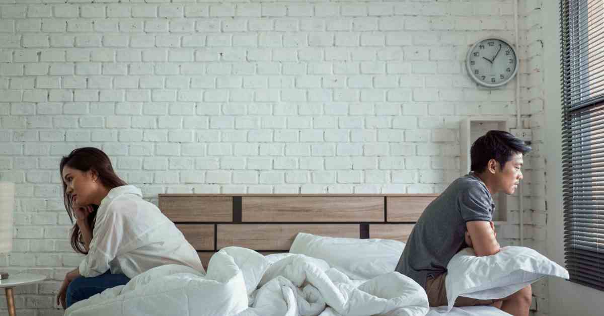 An Asian couple sitting on opposite sides of the bed facing away from each other with sad expressions.