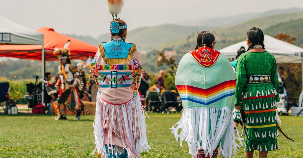 Three women wearing traditional garments with their backs facing the camera are walking into a Chumash Day Pow Wow.