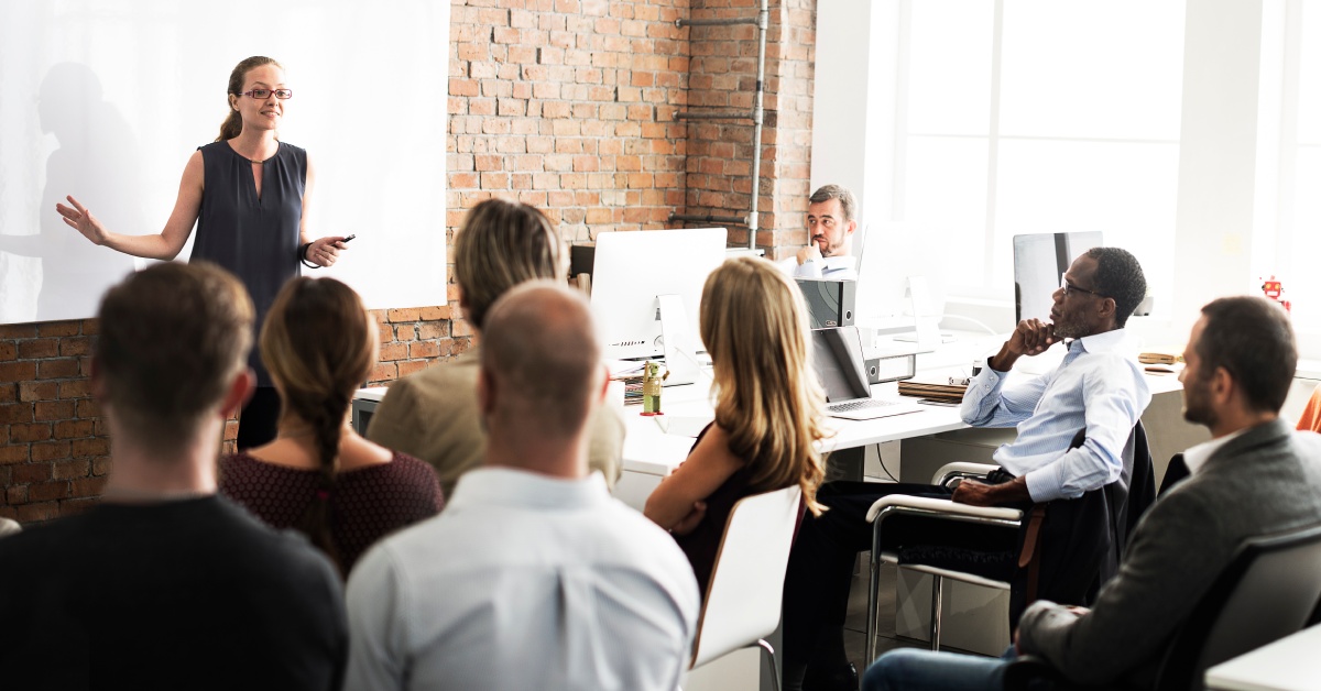 A woman standing in front of a whiteboard talking to a room full of people listening to her training lecture.