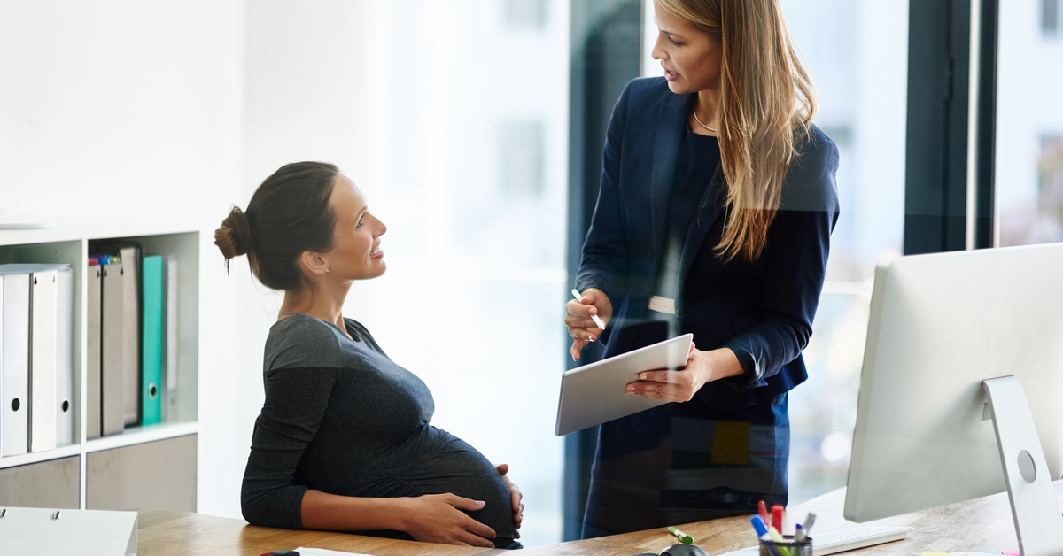 A pregnant woman holding her stomach as she sits at her desk, looking up to talk with one of her female coworkers.