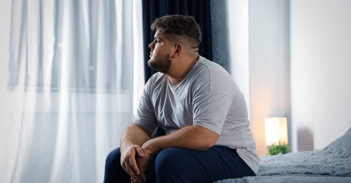 A husky, broad-shouldered man with a beard is sitting on the edge of the bed looking wistfully out the window.