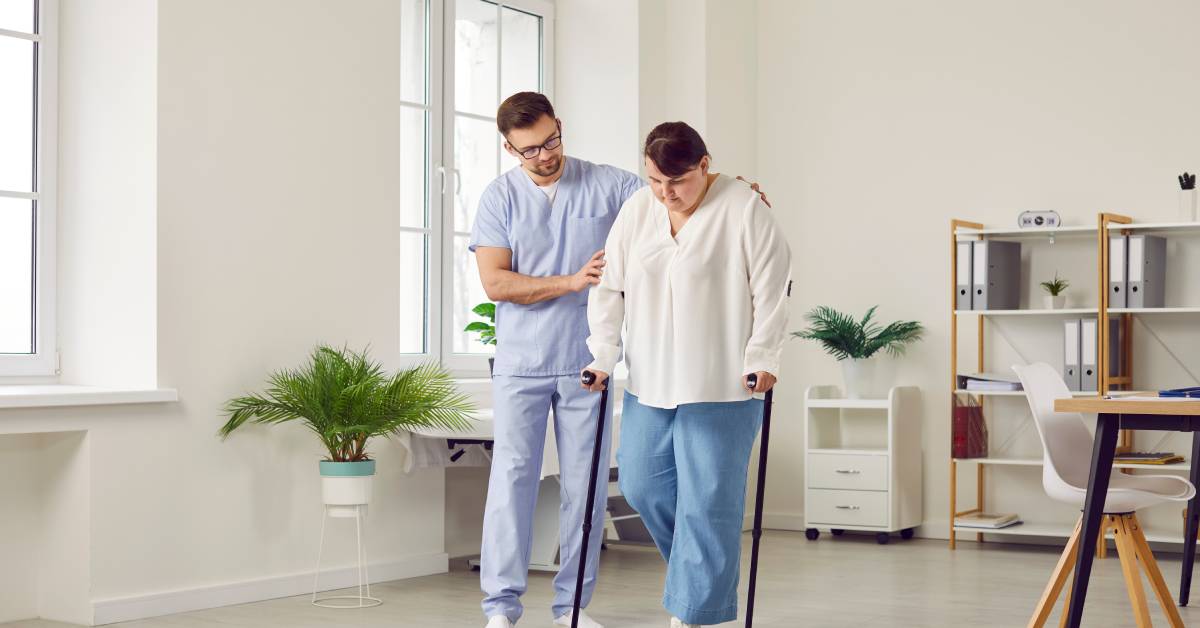 A male nurse in scrubs physically supports a larger woman as she learns to walk on a set of crutches.