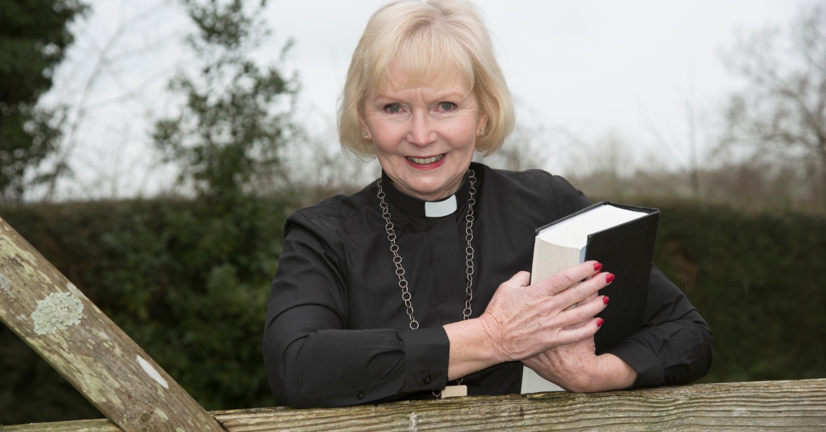 An elderly woman wearing a cassock while smiling and holding a large bible leaning against a garden gate.