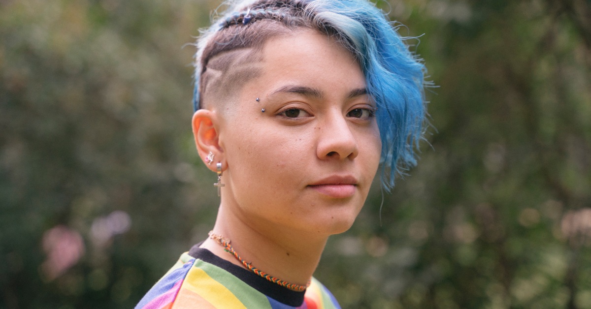 A portrait of a person with blue hair and a side cut wearing multiple piercings and a rainbow shirt.