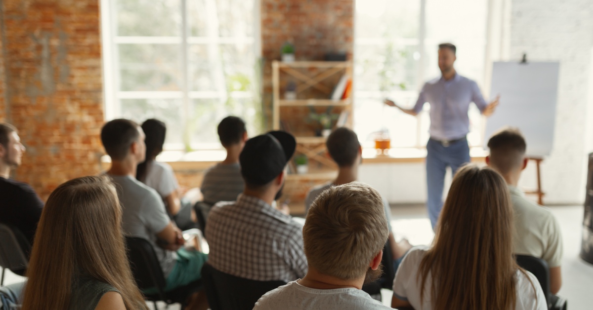 A male speaker at the front of a room blurred out as he speaks to the rest of the room in the foreground.