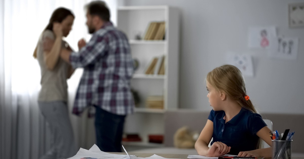 A man becoming violent with a woman in the background while a child in the foreground sits at her desk and watches them.