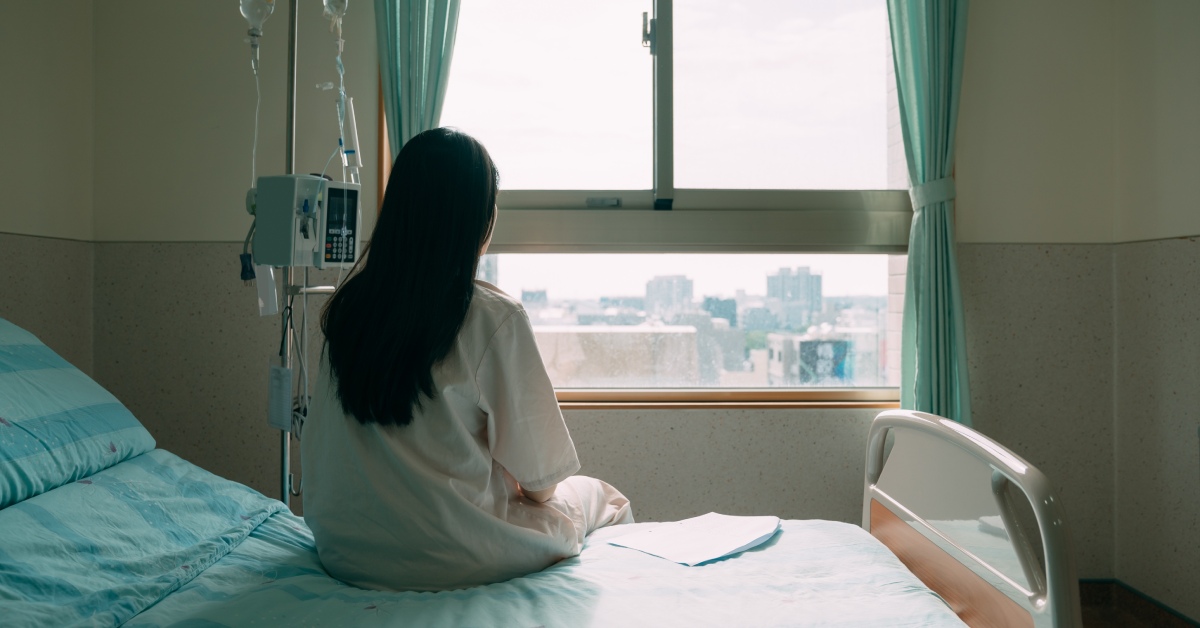 The back of a woman with long black hair sitting on the edge of a hospital bed while looking out the window.