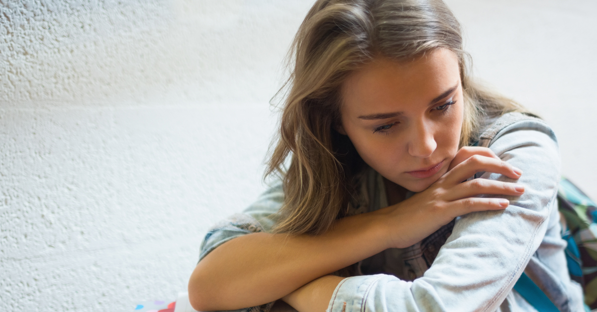 A blonde female student sitting on the stairs holding her binders in her lap as she looks sadly off-screen.