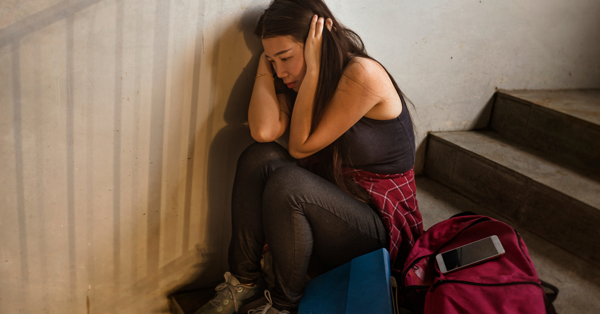 A brunette female student sitting on the stairs while she leans against the wall and covers her ears.