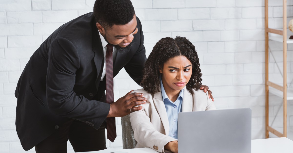 A woman sitting at her desk in front of her computer as she looks uncomfortably at her male coworker in her personal space.