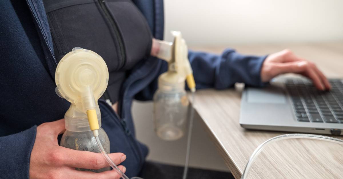 A woman working at her computer desk while holding automatic breastfeeding devices up to her black nursing bra.