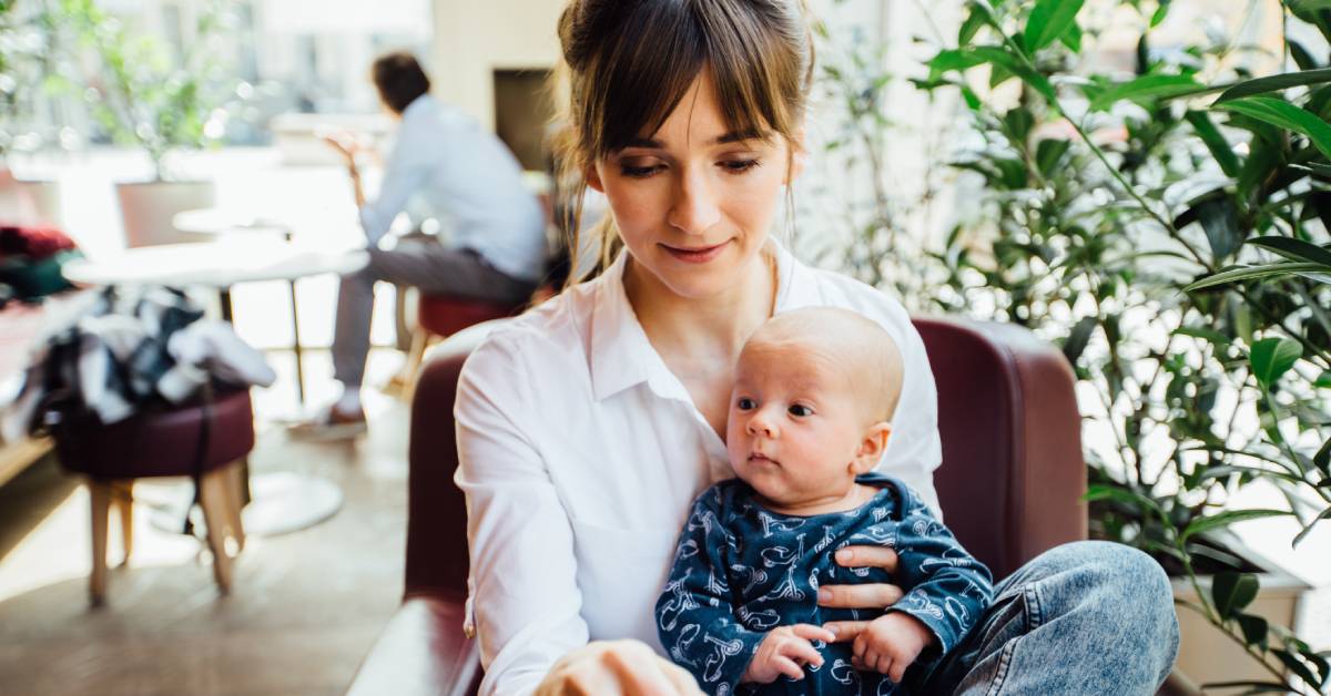 A smiling woman sitting in a comfortable chair at a cafe working on her laptop with her infant on her lap.