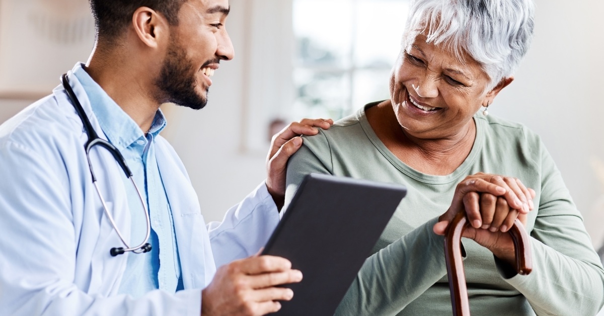 An older female patient and a young male doctor smiling at each other with the doctor's hand placed gently on her shoulder.