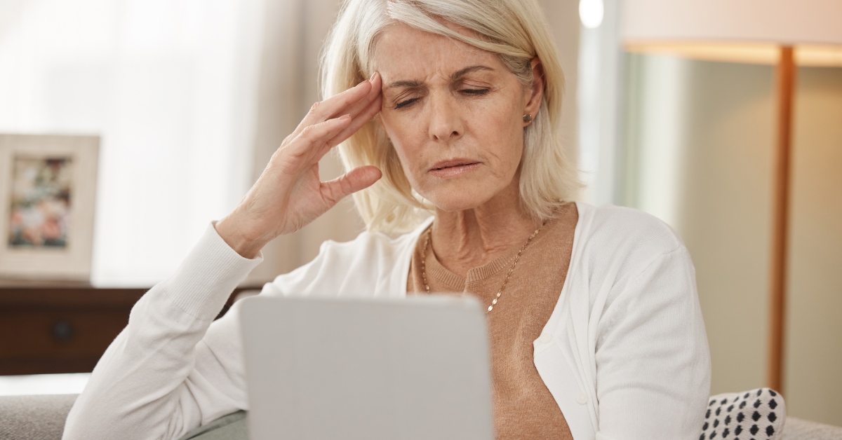 A senior woman sitting on a couch rubbing her temple in pain and frustration as she looks down at her tablet.