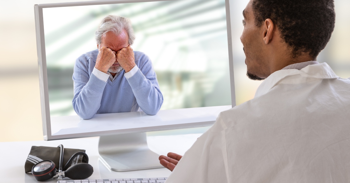 A doctor with a blood pressure cuff on his table, sitting in front of his computer screen, talking to a depressed male senior.