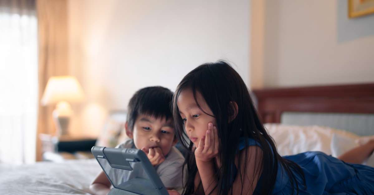 Two young siblings—a boy and a girl—lying down next to each other while they look at a tablet screen.