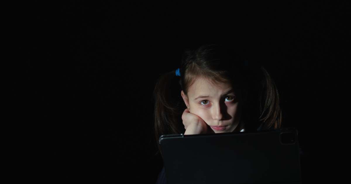 A young girl with pigtails looking sad and tired in front of her tablet in front of a black background.