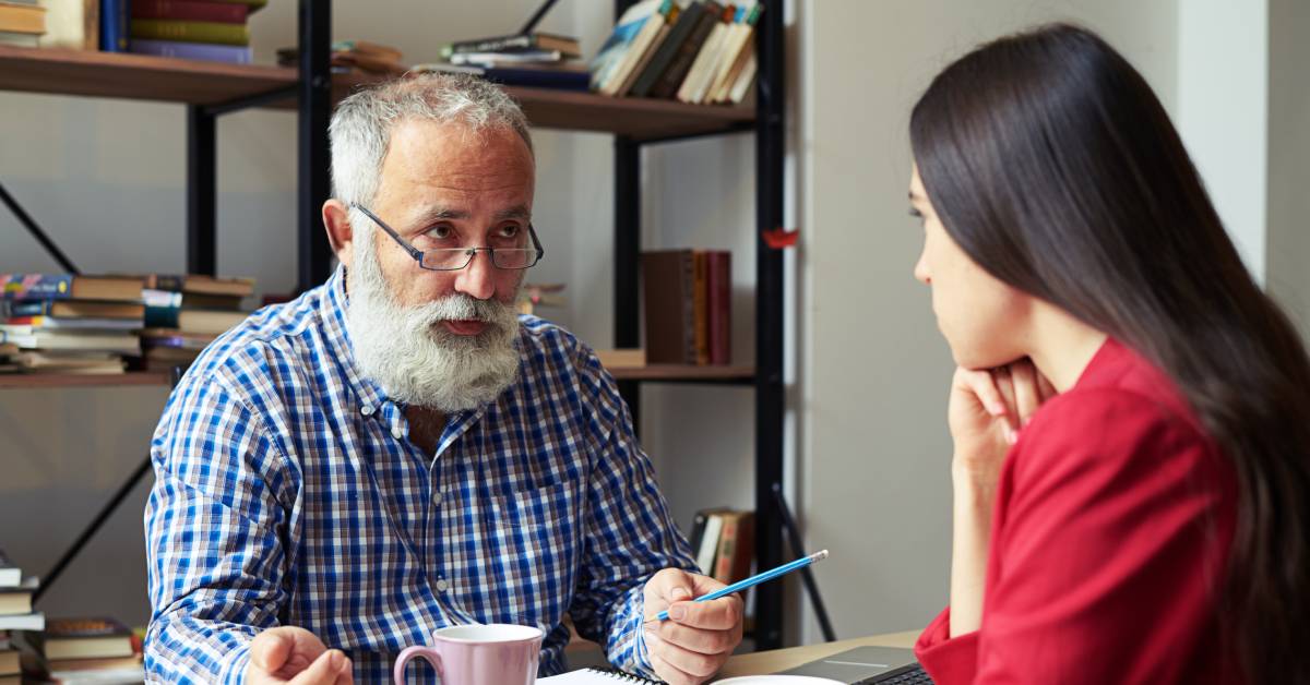 An older man with a beard and glasses, holding a pen, sitting across from a younger woman as they have a serious discussion.