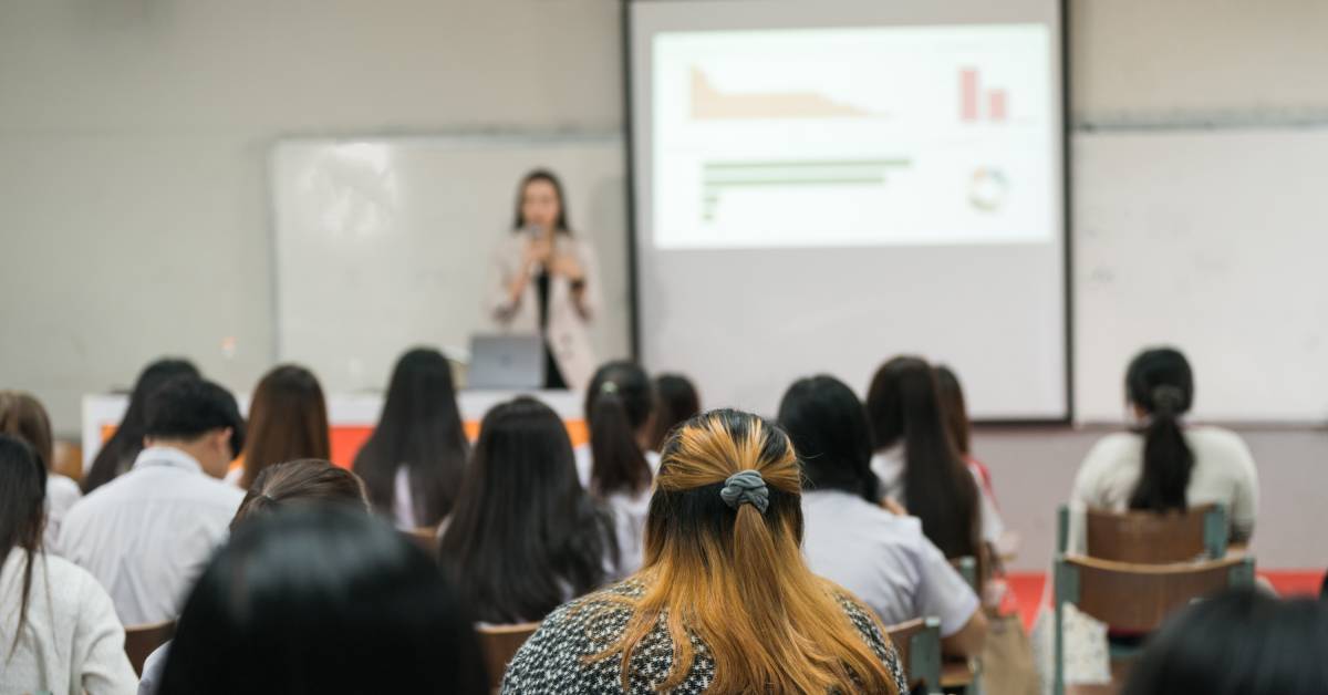 The rear view of a classroom with university students in the foreground and a speaker lecturing at the front of the room.