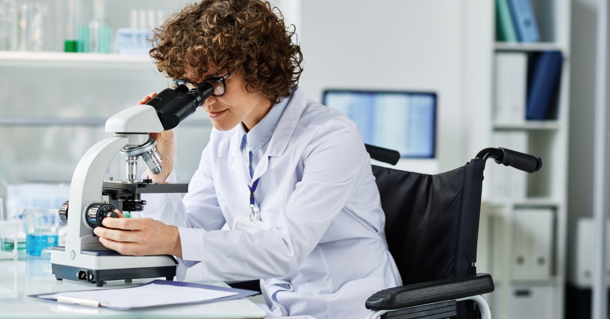 A scientist with curly hair and glasses sitting in their wheelchair looking into the lens of a microscope.