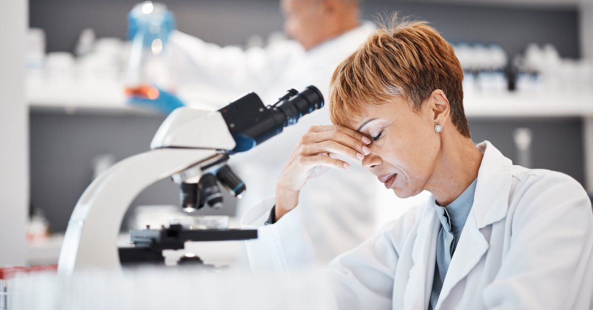 A female scientist sitting at her desk in front of a microscope pinching the bridge of her nose in frustration.