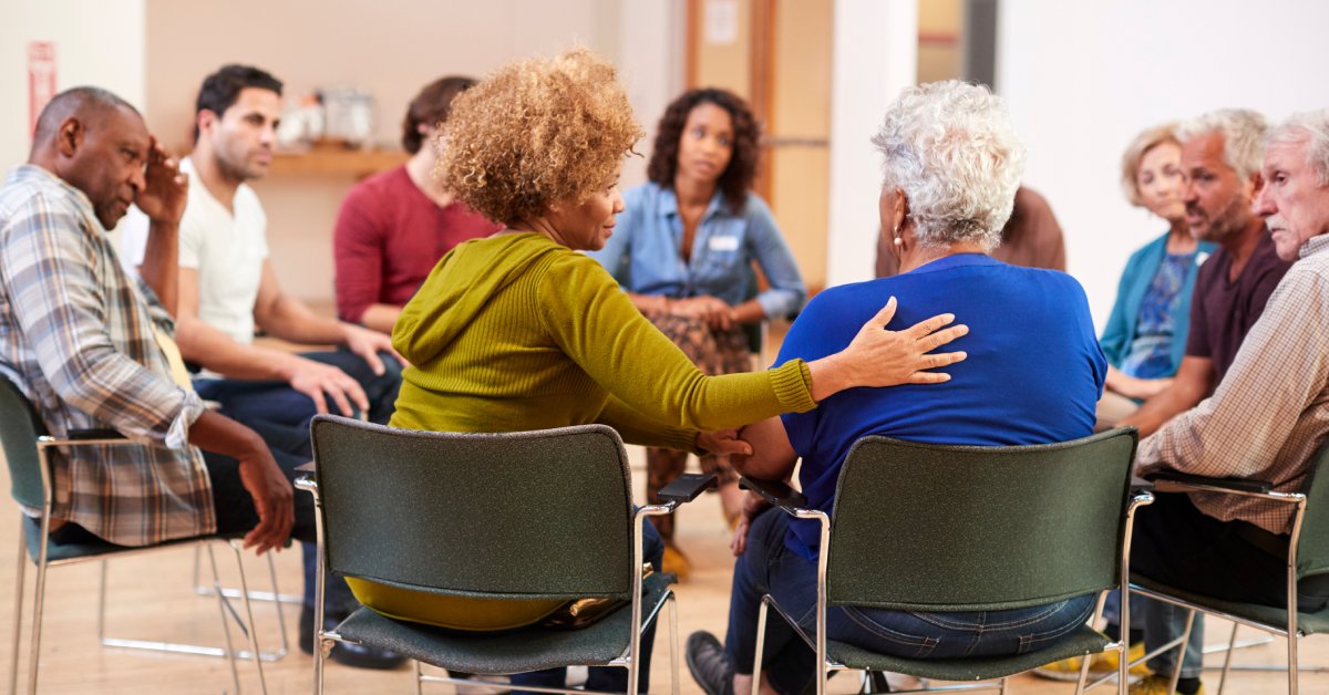 People of different ages and ethnicities sitting in a circle with a woman putting a comforting hand on the woman next to her.