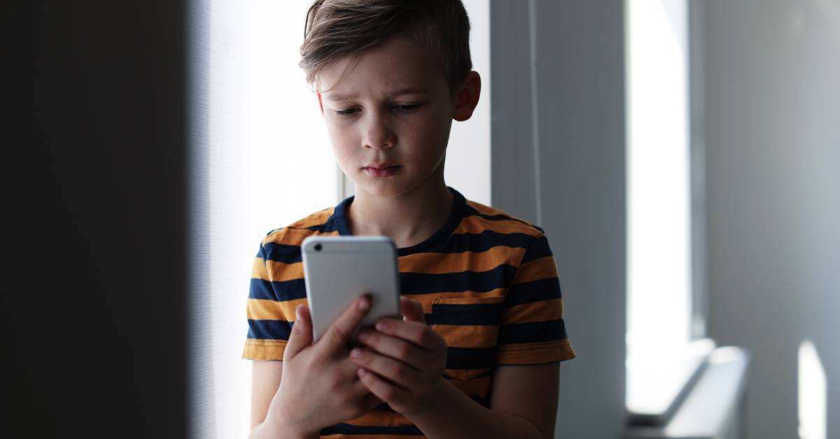 A blonde young boy in a blue and yellow shirt, sitting indoors, sadly looking down at his silver smartphone.