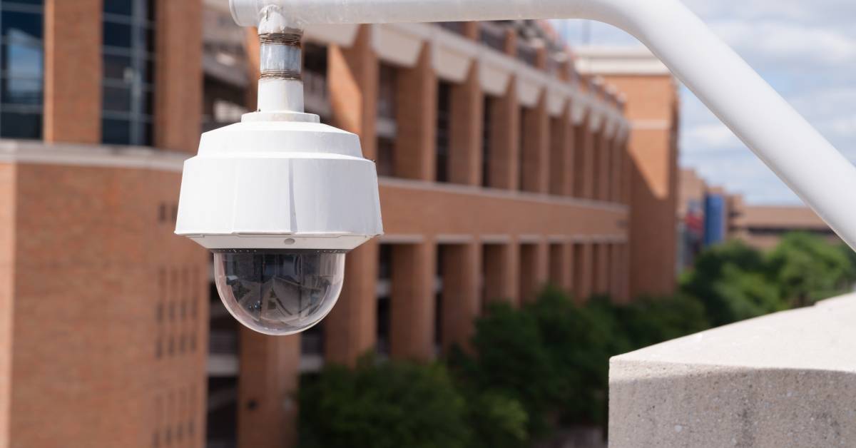 A close-up view of a 360 security camera mounted high above the grounds of a college campus with brick buildings.