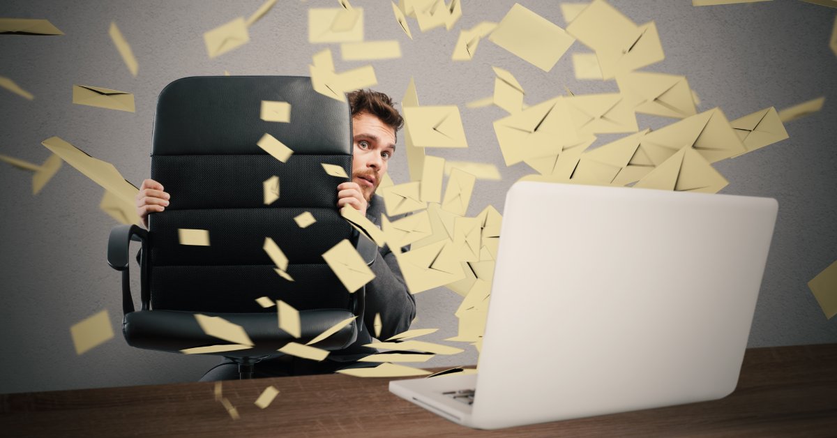 A man crouching down to hide behind his office chair as a flurry of messages bursts out of his computer.