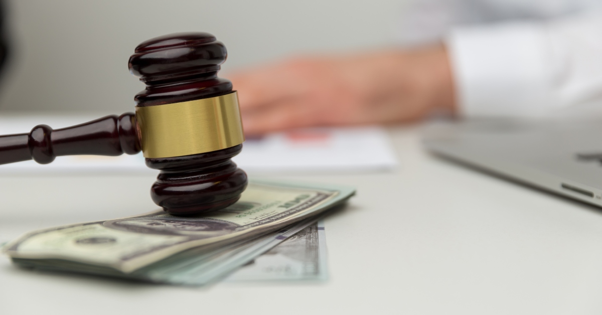 A gavel sitting atop a small stack of money in the foreground and a blurred hand with a white sleeve in the background.