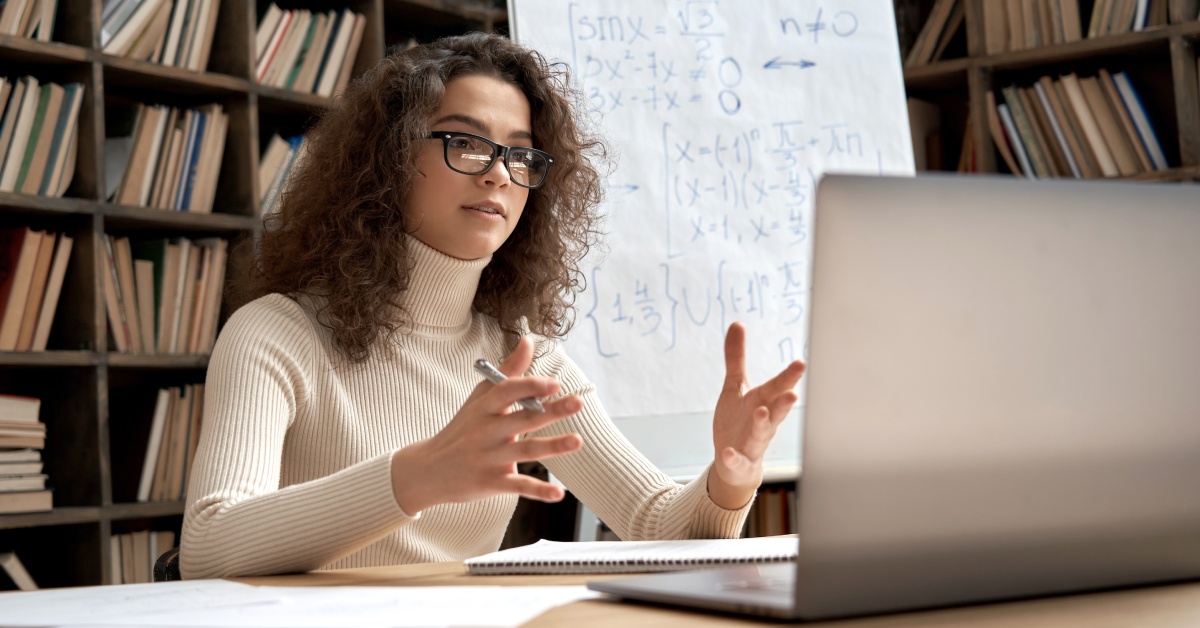 A woman speaking in front of a laptop. A large notepad with equations and a wall of books are in the background.