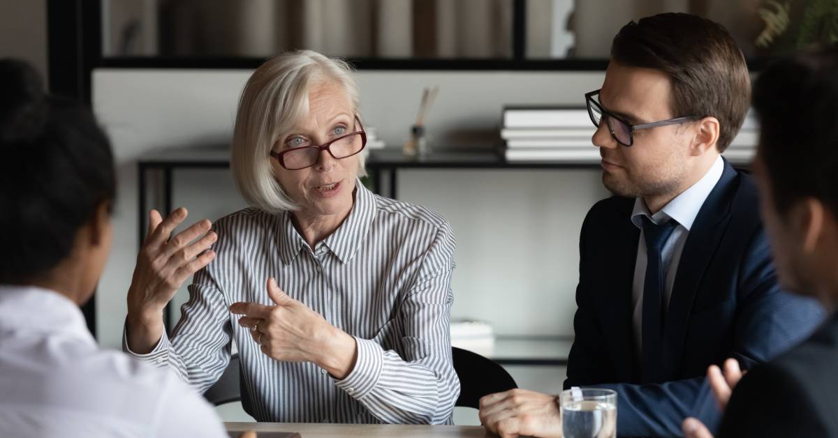 An older woman wearing glasses and gesticulating while speaking to a diverse group of professionally dressed individuals.