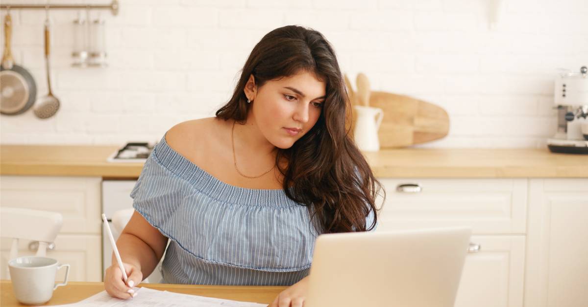 A young woman wearing a halter top, sitting in her kitchen and looking at her computer while taking notes.