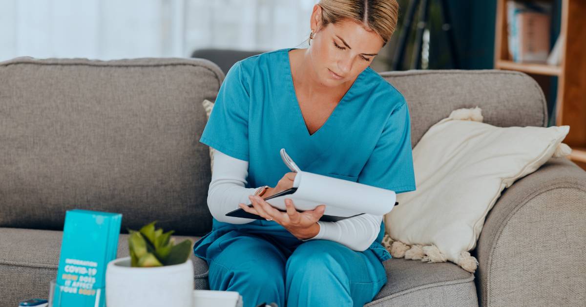 A female nurse in teal scrubs sitting on a grey couch as she writes on a clipboard with a silver pen.