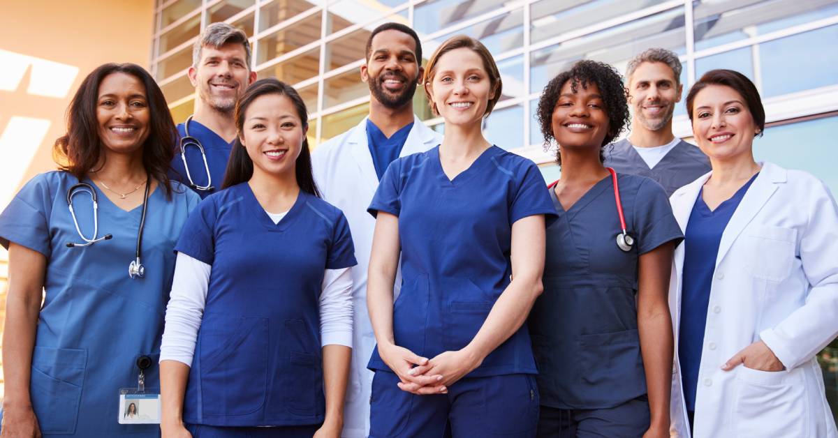 A smiling medical team in dark blue scrubs, white lab coats, and stethoscopes standing outside together.