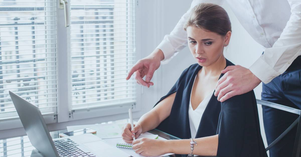 A businesswoman sitting at her computer desk. A man has placed his hand on her shoulder and she stares at it.