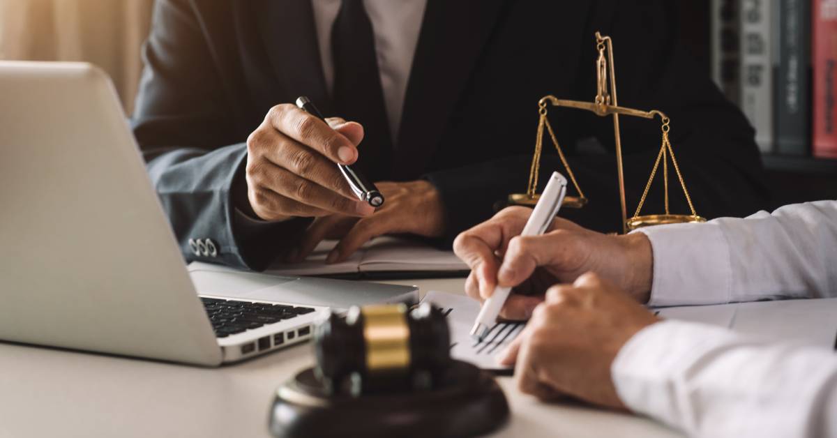 A laptop, a golden scale, and a gavel and sounding block on a table. Two businessmen use pens to gesture to a document.