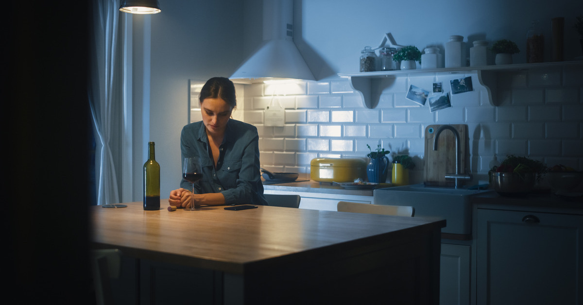 A woman standing alone in her dimly lit kitchen, looking down at the table. An almost empty bottle is next to her.