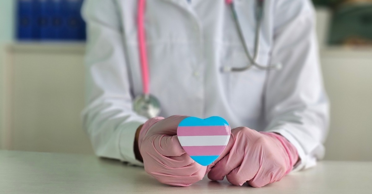 A doctor wearing pink gloves and a pink stethoscope is holding a trans flag in the shape of a heart.