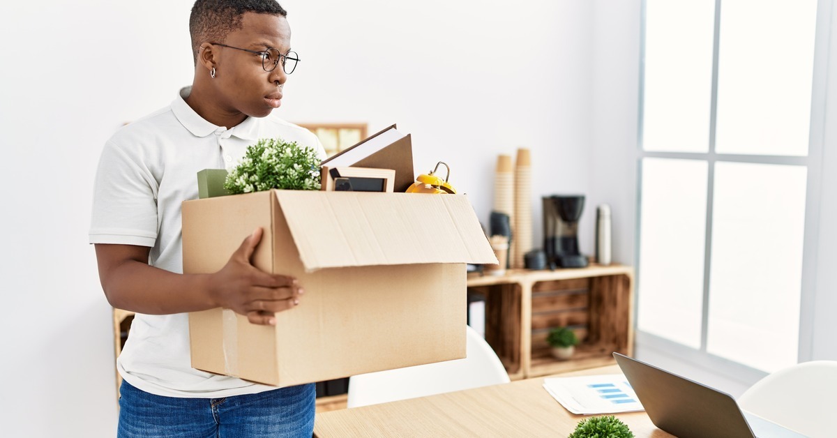 A young, Black adult holding a box of belongings in a white-walled room. They look to the side out the window.