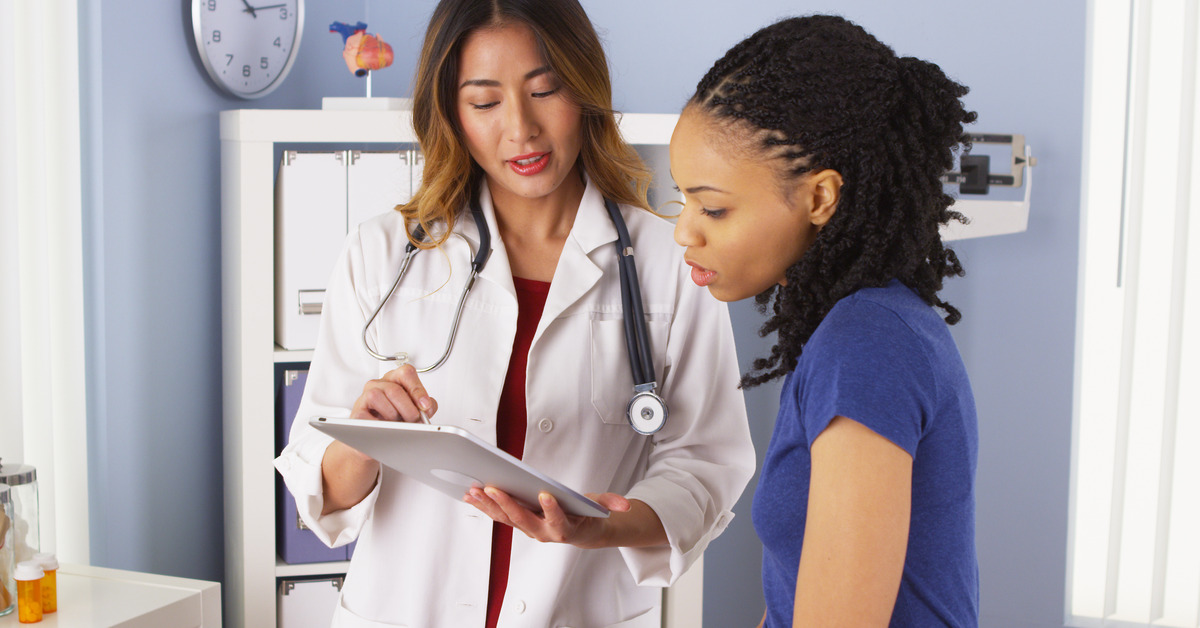 A doctor in a lab with a stethoscope around her neck pointing at the tablet as she talks to her patient.