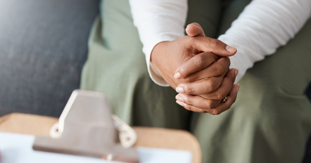 A close-up of a woman's clasped hands wearing a ring. In the foreground, there is a hand taking notes on a clipboard.