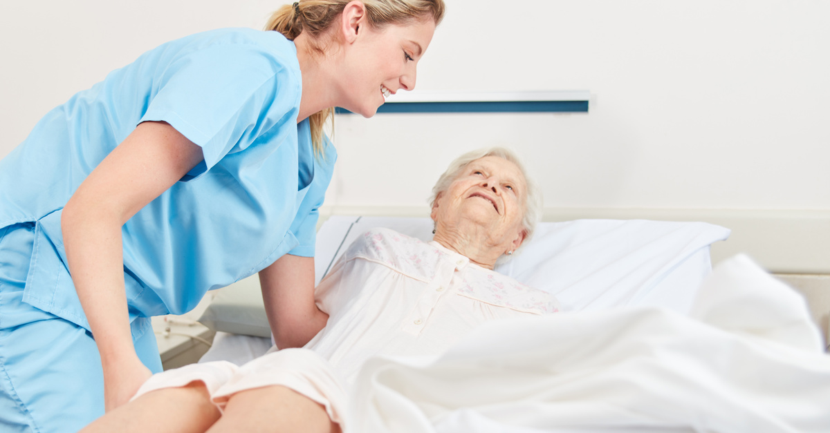 A young female nurse in light blue scrubs lifts and repositions an older woman in her hospital bed.