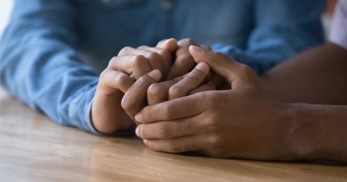 A close-up of two people clasping hands on top of a wood surface. One of the people wears a blue button-up shirt.