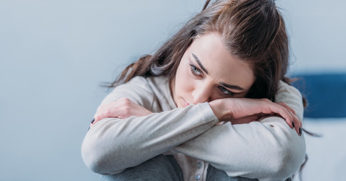 A somber brunette woman wearing gray clothing, hugging her legs with her head on her knees.