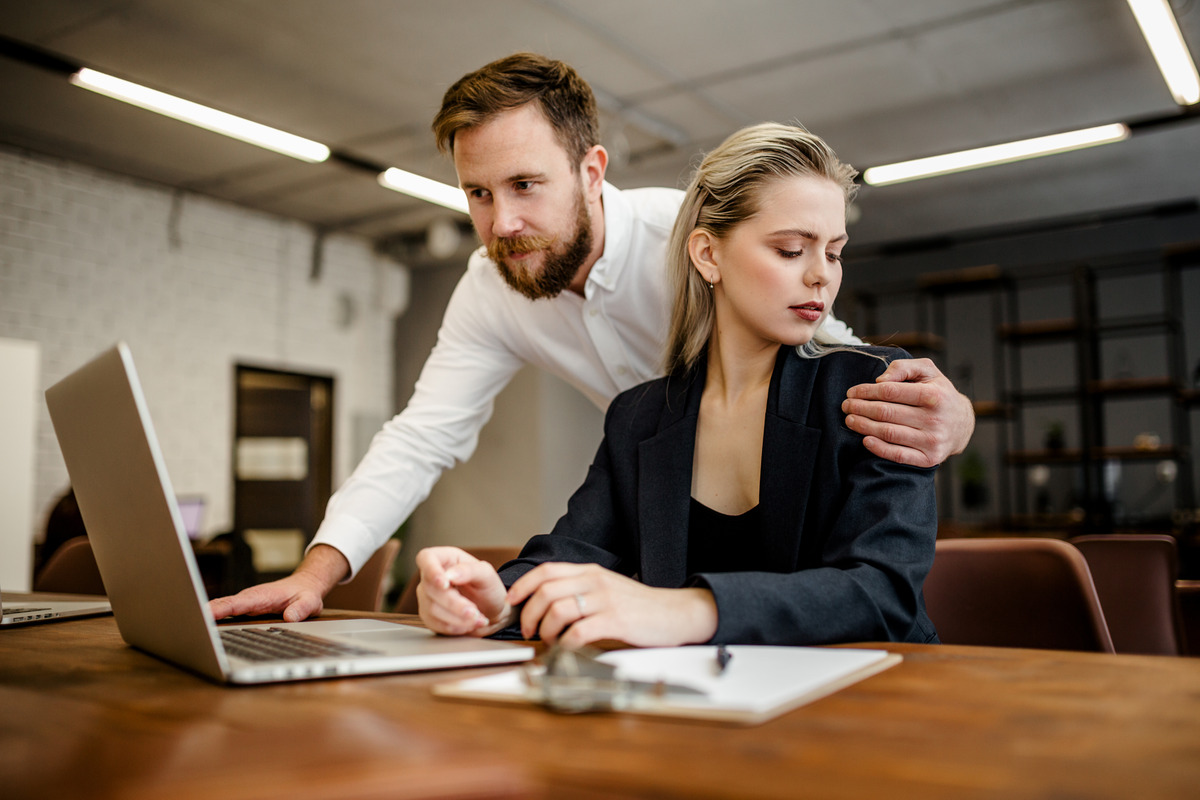 A bearded man leaning over the shoulder of his nicely dressed co-worker as she looks uncomfortably at his hand.