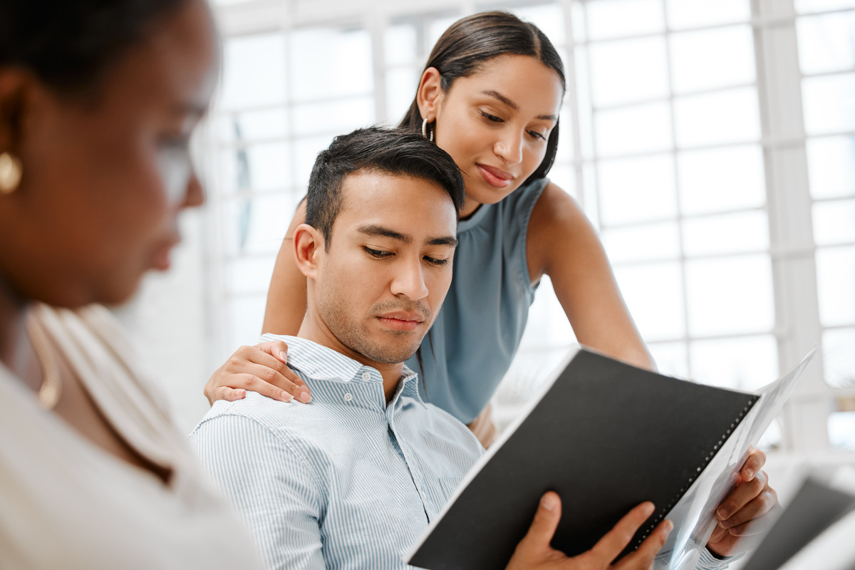 A woman showing her co-worker something in an open folder. She touches his shoulder. He looks uncomfortable.